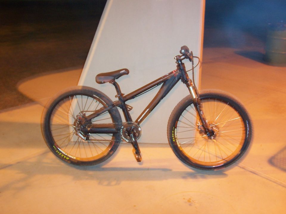 Black hardtail dirt jump mountain bike with Maxxis tires leaning on a white wall at night