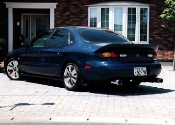 1999 Ford Taurus - A blue 1999 Ford Taurus sedan is parked outside a brick building, viewed from the rear.
