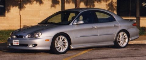 1999 Ford Taurus - A silver four-door Ford Taurus sedan photographed from a front three-quarter angle, showing multi-spoke alloy wheels, body-side molding, a…