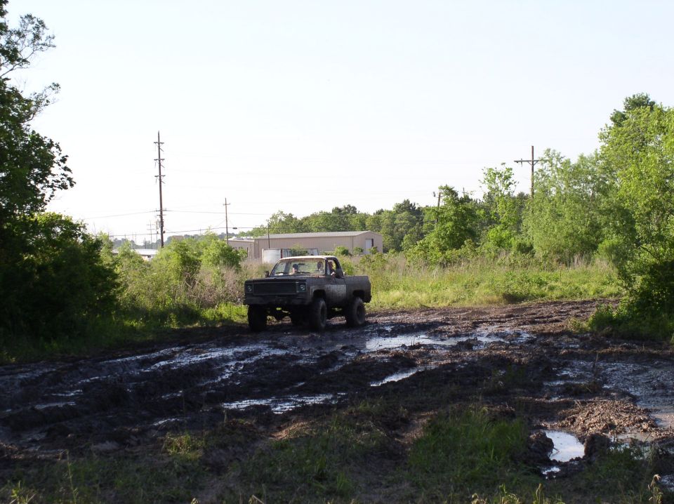 Old pickup truck with square body and aftermarket grille driving through a muddy track. Trees and buildings in background