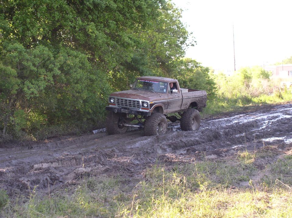 Brown lifted pickup truck driving through a muddy track with trees on the left and a bright sky on the right