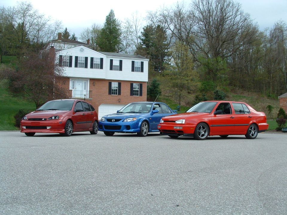 Red sedan, blue sedan, and red hatchback parked on asphalt in front of a brick and white house