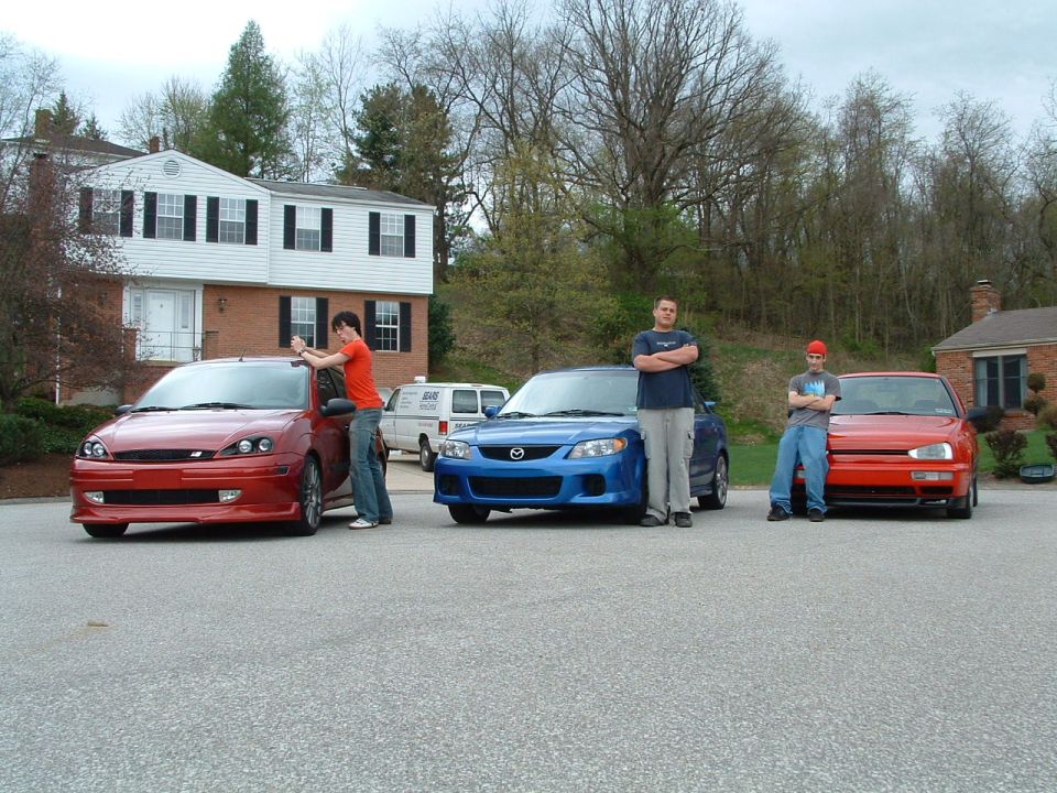 Red Ford Focus, blue Mazda, and red Volkswagen parked in front of houses with people standing by them