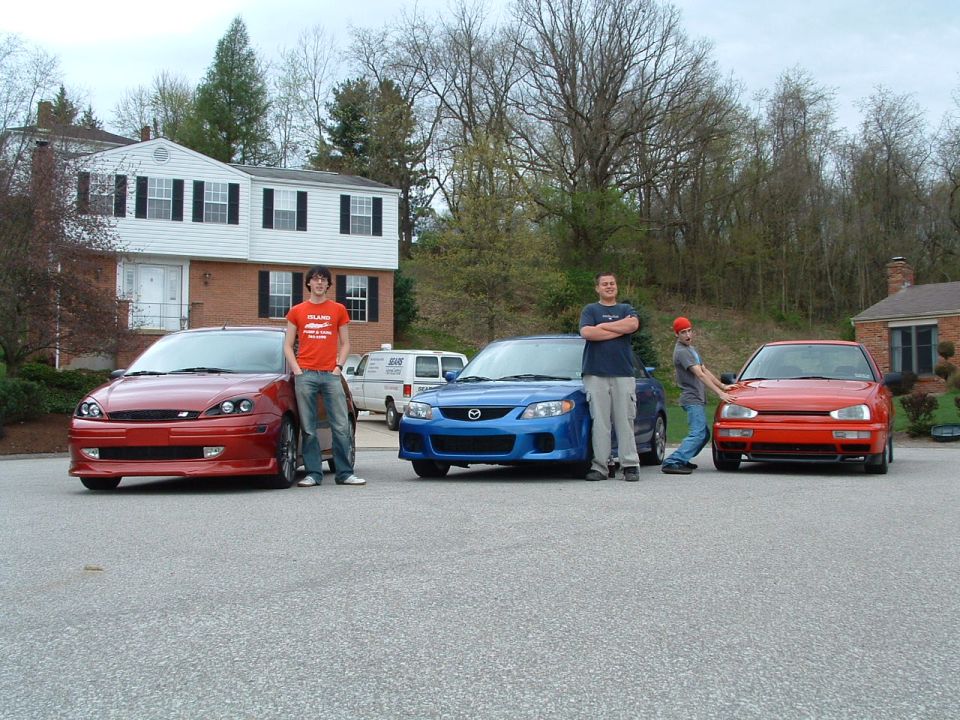 red Ford Focus, blue Mazda 3, and red VW Golf parked in driveway with people