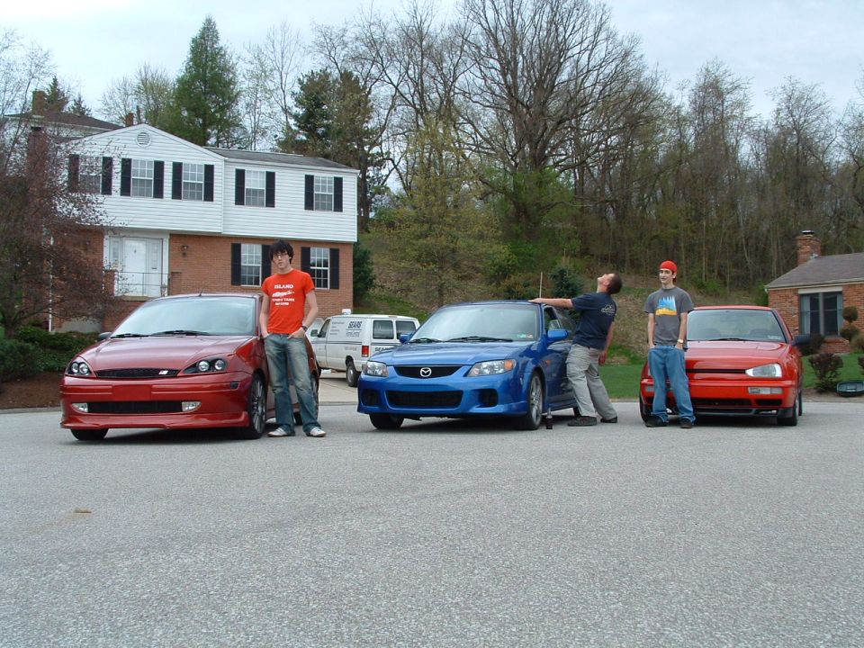 Red Ford Focus, blue Mazda 3, and red Volkswagen Golf parked in a driveway with people