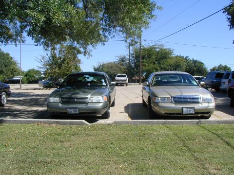 2000 Ford Crown Victoria - Two Ford Crown Victorias are parked in a lot under a clear sky with trees in the background.