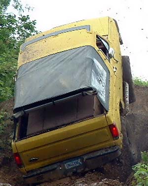 1988 Ford Bronco - A yellow Ford Bronco is seen tilted on its side on a dirt incline, with its rear facing the camera.