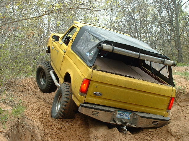 Yellow Ford Bronco, top off, rear 3q view, climbing sandy wooded trail on mud tires