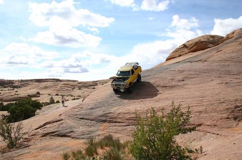 1988 Ford Bronco - Yellow off-road vehicle navigates a steep, rocky desert landscape under a partly cloudy sky.