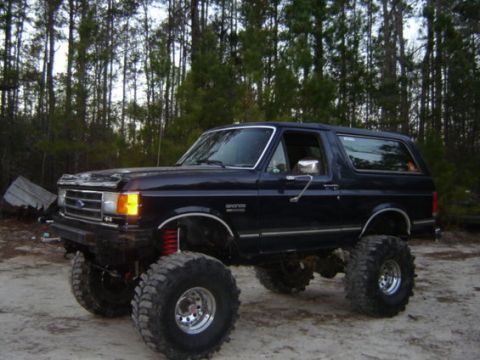 1978 Ford Bronco - A lifted black Ford Bronco sits on a dirt lot with trees in the background.