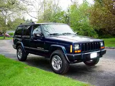 1988 Jeep Cherokee - A dark blue Jeep Cherokee is parked on a paved surface with green grass and trees in the background.