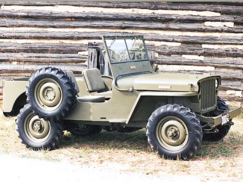 1988 Jeep Cherokee - This vintage Jeep, likely a CJ model, is presented in a utilitarian olive drab finish against a rustic log cabin backdrop.