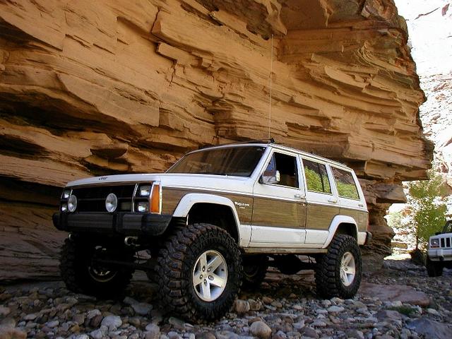 White Jeep Wagoneer with woodgrain sides, lifted, on rocks under a sandstone overhang