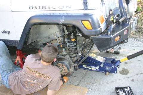 2003 Jeep TJ - A mechanic works on the suspension of a white Jeep TJ, which is supported by a jack and jack stands.