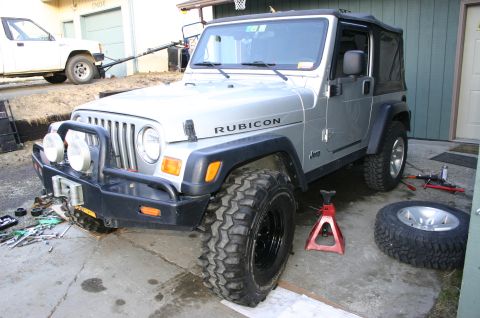 2003 Jeep TJ - A silver 2003 Jeep TJ Rubicon is parked in a driveway, undergoing tire maintenance with a jack and spare tire visible.