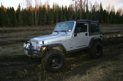 2003 Jeep TJ - This silver Jeep Wrangler features an off-road lift and Rubicon badging, seen navigating a muddy, wooded area.