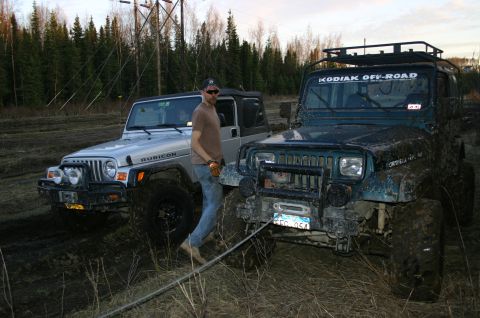 2003 Jeep TJ - Two off-road Jeeps are bogged down in mud, with a man standing between them. One Jeep has a winch and a roof rack.