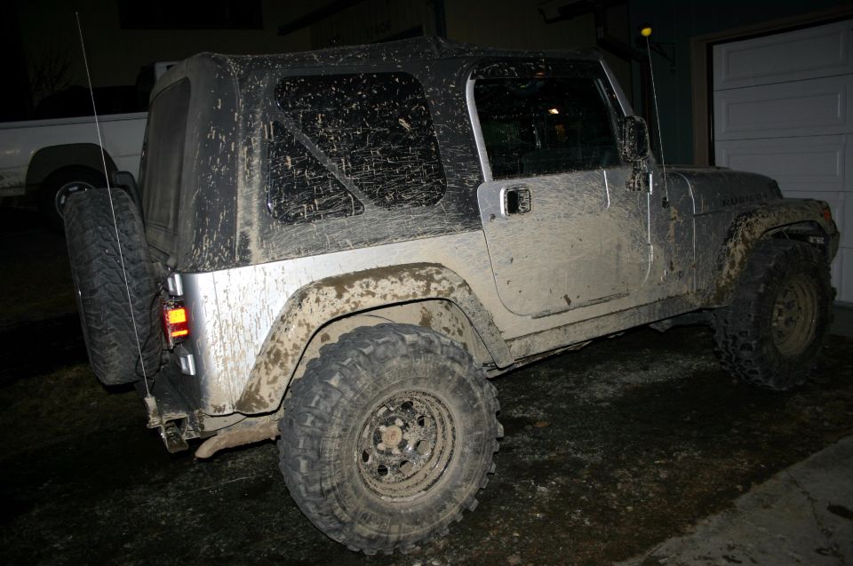 Muddy Jeep Wrangler parked in front of a garage. It has a soft top and a spare tire on the back