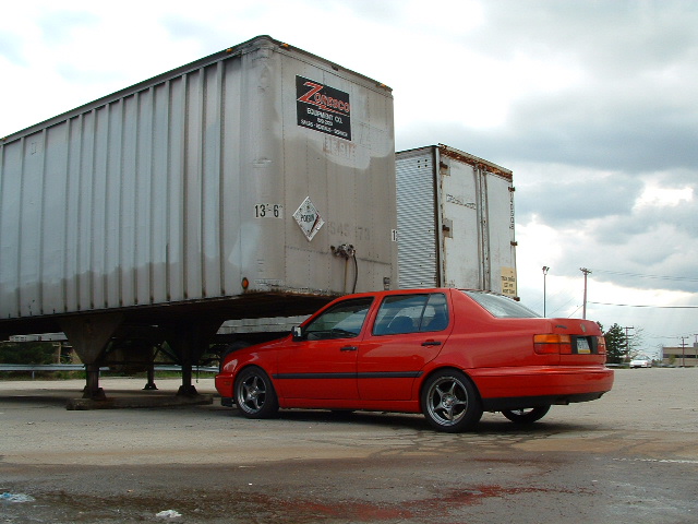 Red sedan, possibly a Volkswagen Jetta, parked beside a large trailer. The car has aftermarket wheels and appears to be from the early to mid-1990s