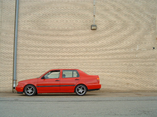 1994 Volkswagen Jetta sedan, red, parked on asphalt facing a tan brick wall