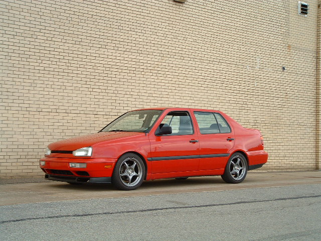 Red sedan parked in front of a brick wall. It has aftermarket wheels