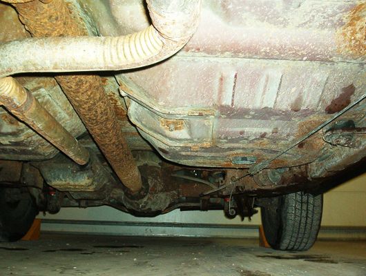 Rusty undercarriage of a car showing corroded exhaust, fuel tank, and rear suspension in a garage