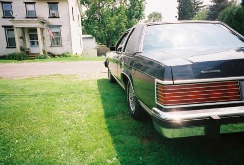 1984 Mercury Grand Marquis - Rear view of a dark sedan parked on a lawn, with a white house in the background.