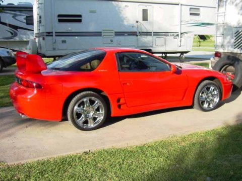 1999 Mitsubishi 3000GT - A red sports car with a distinctive rear spoiler is parked on a concrete surface.