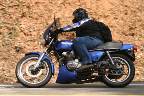 1985 Ford LTD Crown Victoria - A rider on a blue Suzuki motorcycle travels down a road with a wooded embankment.