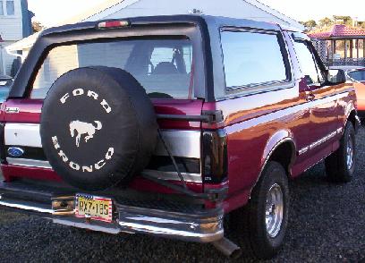1995 Ford Bronco - Rear three-quarter view of a red Ford Bronco with a black hardtop, parked on gravel in daylight. A swing-away carrier holds the spare under…