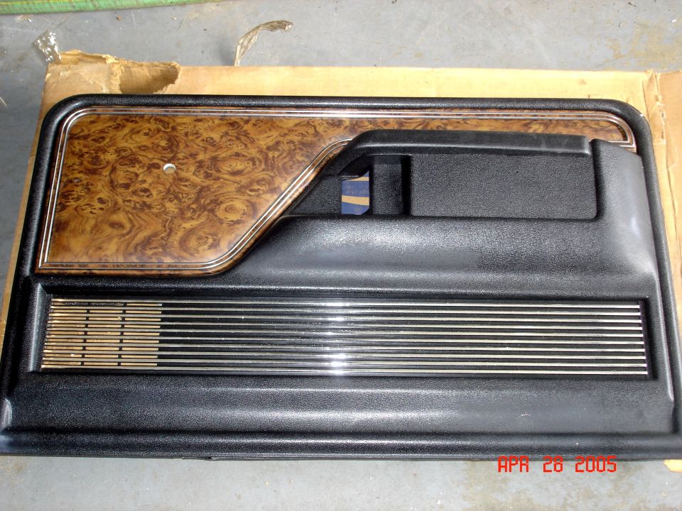 Black interior door panel with woodgrain insert and chrome accent strips lying in a cardboard box