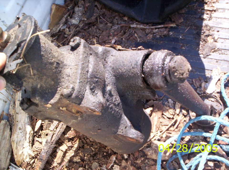 Black, greasy steering gear box with a metal rod extending from the right side, resting on wood and debris
