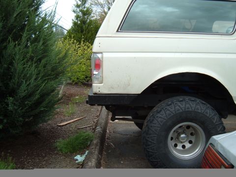 1989 Ford Bronco - The rear of a white SUV is visible, parked next to a curb with trees in the background.