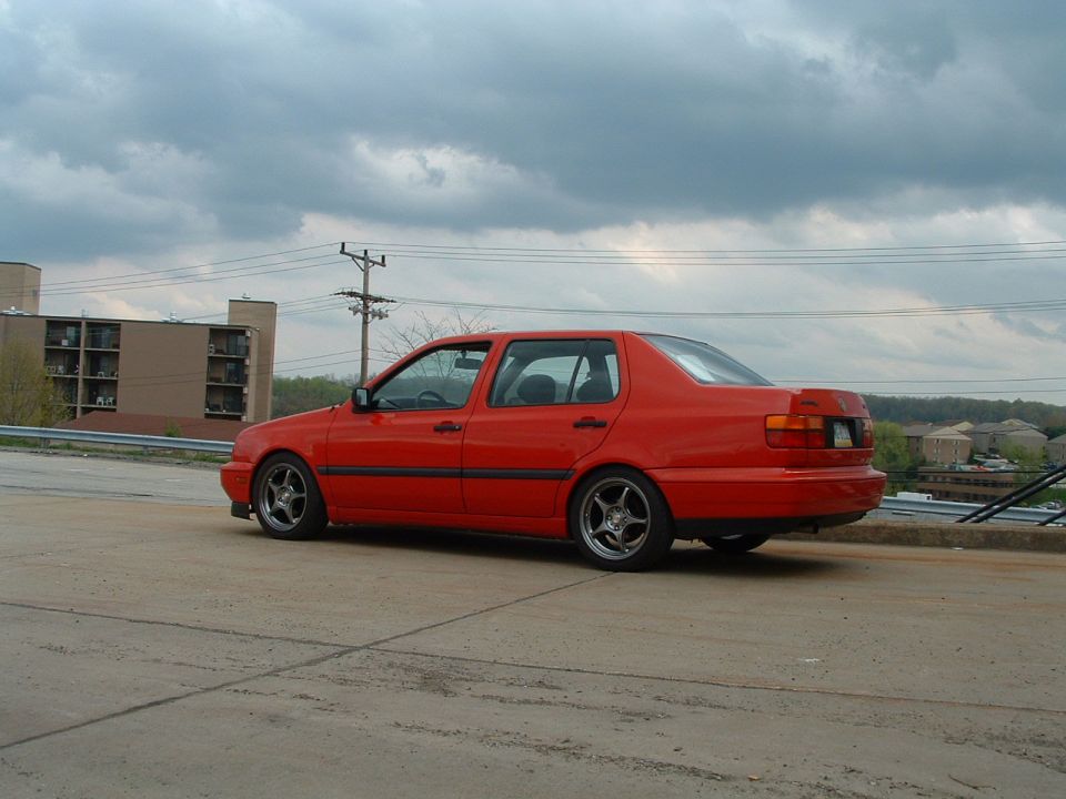 Red sedan parked on concrete, side view. Aftermarket wheels, slight body modifications
