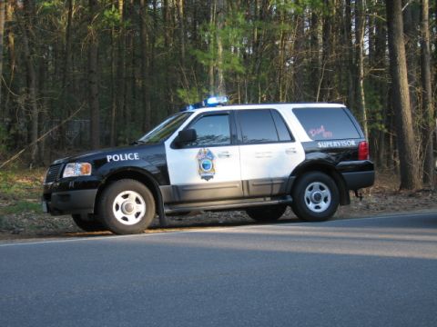 1991 Ford LTD Crown Victoria - A police SUV with flashing lights is parked on the shoulder of a road, with a wooded area behind it.