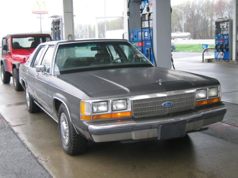 1991 Ford LTD Crown Victoria - Gray Ford Crown Victoria sedan at a gas pump island on a wet overcast day, shot from the 3/4 front right with parking lights on. Its hood a…