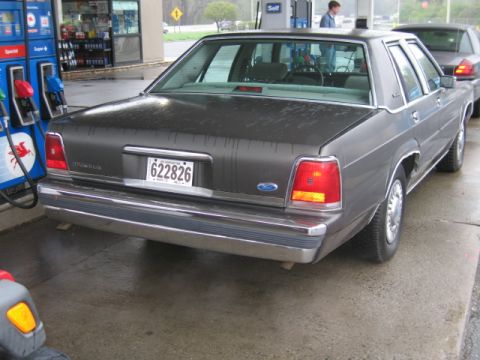 1991 Ford LTD Crown Victoria - Gray Ford Crown Victoria sedan seen from the 3/4 rear left at a self-serve gas station on a wet day, with pumps and the station canopy behi…