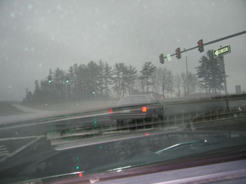 1991 Ford LTD Crown Victoria - A dark sedan drives through a rainy intersection, with traffic lights and bare trees visible in the background.
