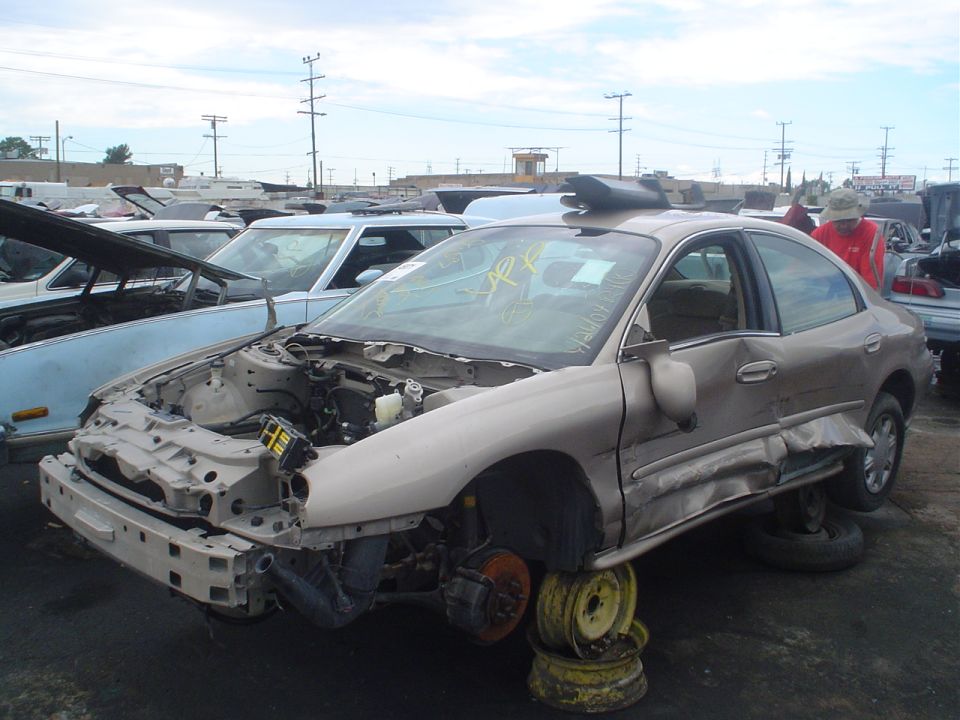 Wrecked sedan with front end damage, missing hood and bumper, propped on rims in a junkyard. Another car is visible to the left