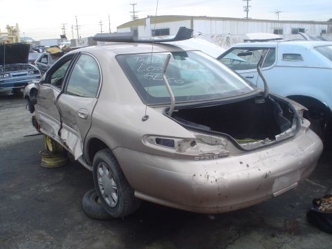 1993 Ford Taurus - A damaged tan sedan sits in a junkyard with its trunk open and rear quarter panel severely dented.