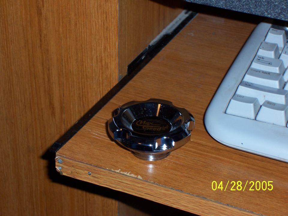 Chrome gas cap with "Max Speed" text on a wooden surface next to a keyboard
