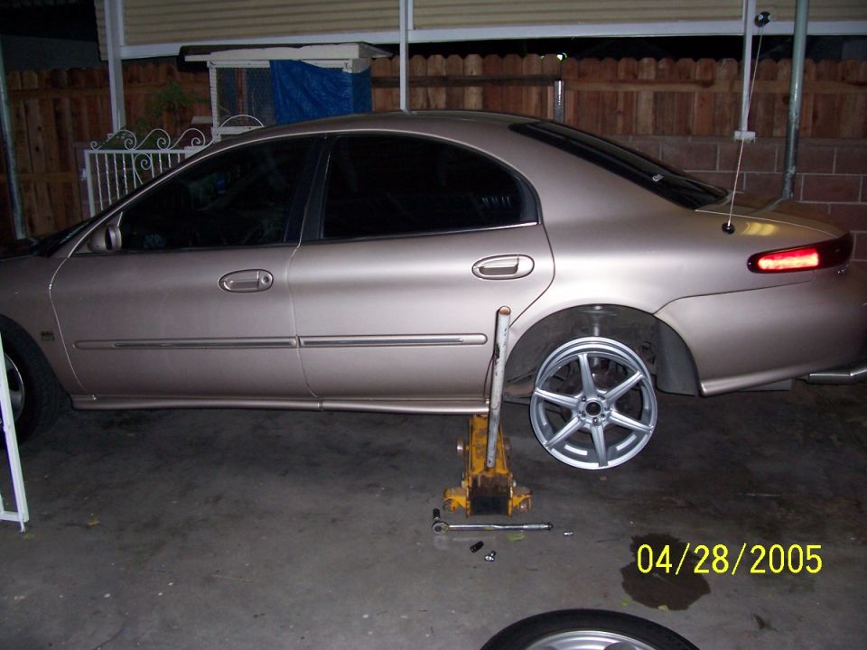 1998 Mercury Sable on a jack with the rear wheel removed, showing a new aftermarket wheel nearby