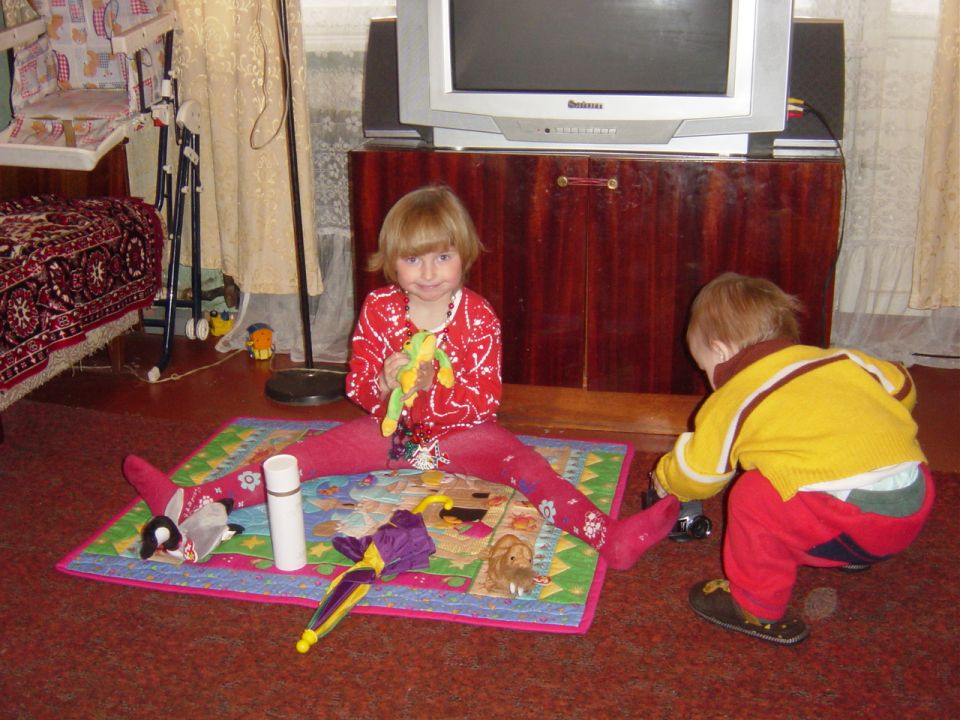 Two young children play on a rug in a room with a television and furniture