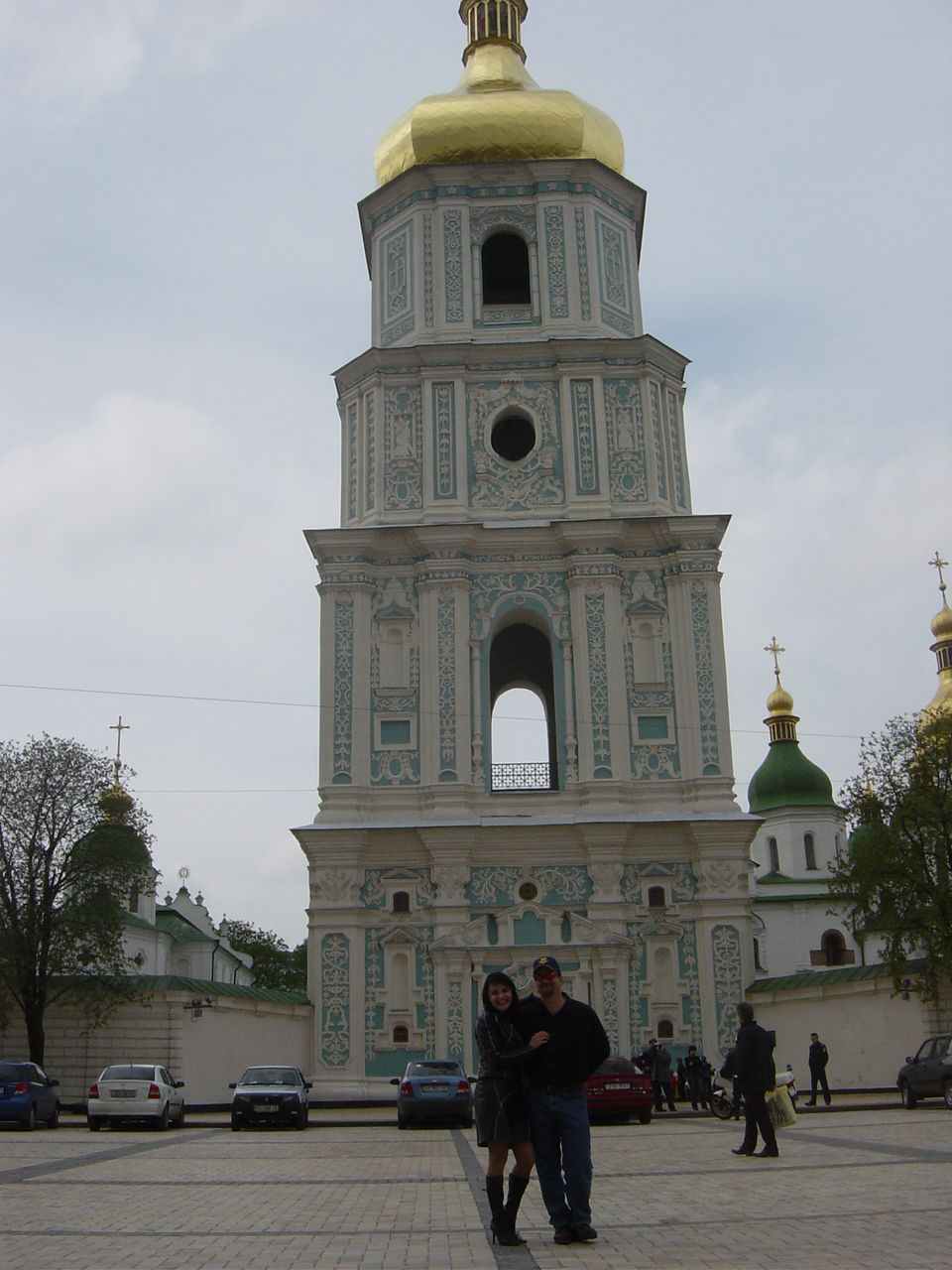 Couple posing in front of a large, ornate bell tower with golden domes. Cars parked in the background