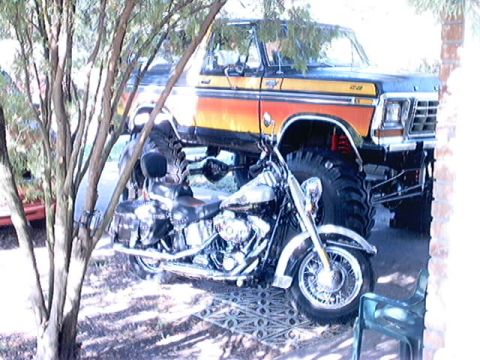 1979 Ford Bronco - A lifted 1979 Ford Bronco with prominent orange and yellow stripes is parked next to a motorcycle, partially obscured by trees.