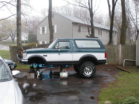 1994 Ford Bronco - A two-tone Ford Bronco is elevated on jack stands in a driveway, undergoing maintenance.