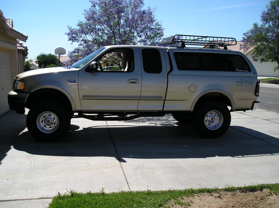 1999 Ford F-150 pickup truck, lifted, with a silver camper shell and roof rack, parked on a driveway