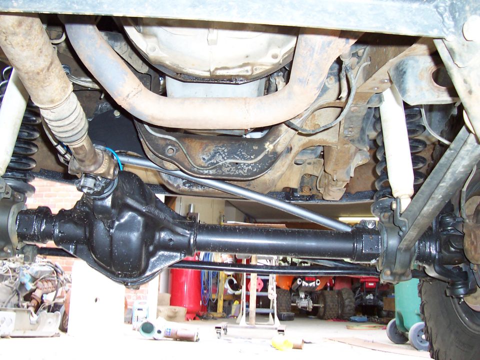 Underside view of a truck's rear axle, driveshaft, and suspension components. Other vehicles are visible in the background