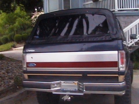 1991 Ford Bronco - Rear view of a lifted dark blue 1980s-90s Ford Bronco fitted with a black soft top over the rear section, parked in a residential driveway.…