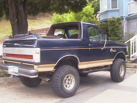 1991 Ford Bronco - Dark blue two-door Ford Bronco SUV with tan lower body accents, viewed from the rear three-quarter angle with the soft top removed. It sits…